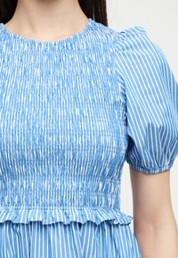Femme portant un top à rayures verticales bleu et blanc avec des manches courtes bouffantes et un corsage smocké avec un ourlet à volants.