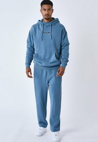 Sudadera azul de algodón con un bolsillo tipo canguro en el frente, puños ajustados y un logo. Combinada con pantalones azules a juego y zapatillas blancas.