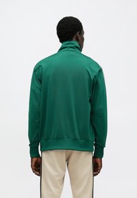 Grønn atletisk jakke med glidelås, høy krage, glatt tekstur og elastiske mansjetter. Har subtile designdetaljer langs skuldrene.