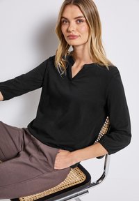 Zwarte langslavige top met een ingesneden kraag, relaxed fit en zachte textuur, gecombineerd met lichtbruine broek met elastische taille.