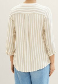 Personne portant un chemisier à manches longues beige et blanc à rayures verticales avec un détail plissé dans le dos, associé à un jean en denim bleu clair.