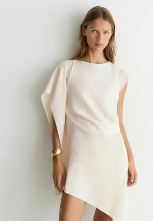 Frau trägt ein elfenbeinfarbenes asymmetrisches Kleid mit drapiertem Ärmel und einem skulpturalen goldenen Armreif, steht vor einer schlichten weißen Wand.