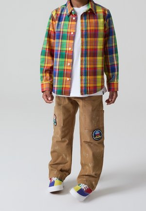 Enfant portant une chemise à carreaux multicolore boutonnée, un t-shirt blanc, un pantalon marron avec des patchs et des baskets multicolores, debout devant un fond gris.