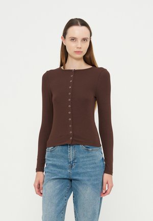 Femme aux longs cheveux bruns portant un haut marron cintré à manches longues avec des boutons sur le devant et un jean en denim bleu, debout devant un fond uni.