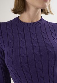 Lila gestrickter Pullover mit einem Zopfmuster, geripptem Ausschnitt und figurnaher Passform, der eine strukturierte Oberfläche hervorhebt.