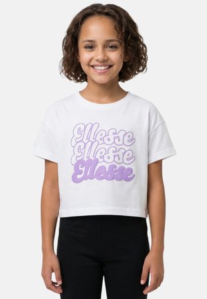 Fille aux cheveux bouclés sourit, portant un t-shirt court blanc avec le texte violet « Ellesse » et un pantalon noir, debout devant un fond clair uni.