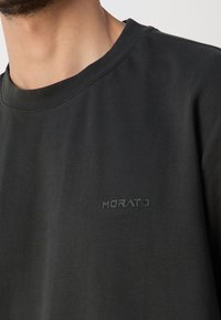 Muž v černém tričku s kulatým výstřihem, na kterém je na hrudi malý černý potisk s logem "MORATO".