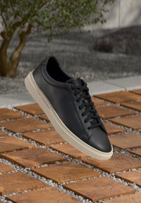 Svarte lær-sneakers med teksturert overflate, rund tå, tonale snøringer og en lys beige gummisåle, svevende over en mursteinsoverflate.