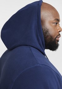 Felpa con cappuccio in fleece blu navy, caratterizzata da un cordoncino, un ampio cappuccio e tessuto testurizzato. La vista laterale ravvicinata mette in risalto i dettagli del materiale e del design.