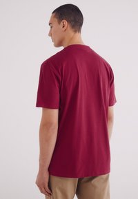 T-shirt in cotone bordeaux con colletto rotondo, maniche corte e vestibilità morbida, vista da dietro, abbinata a pantaloni beige.