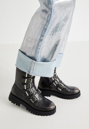 Czarne skórzane botki za kostkę z czterema srebrnymi klamrami i grubą podeszwą z protektorami, noszone z podwiniętymi jasnoniebieskimi dżinsami.