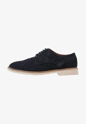 Scarpa brogue in suede nero con design a punta alata, dettagli traforati, lacci neri e suola di colore marrone chiaro.