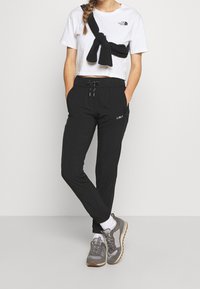 Pantalons de sport noirs avec ceinture élastique et poches, associés à un t-shirt court blanc. Des baskets grises complètent la tenue.