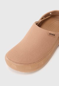 Beige slip-on Crocs clogs med texturerat tygöverdel och gummisula, som har en justerbar rem och en liten tyglogotagg.