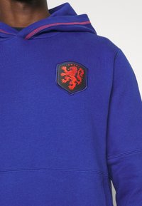Persona che indossa un felpa blu con simbolo di un leone rosso e testo "KNVB" sul petto, mostrando dettagli della parte superiore del corpo e del cappuccio.