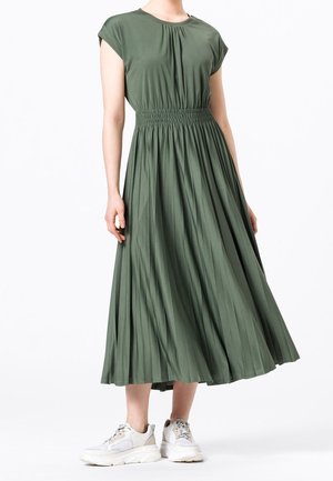 Femme portant une robe plissée olive verte, longueur cheville, à manches courtes, avec des baskets blanches épaisses, sur un fond blanc uni.