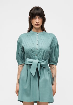 Kobieta ubrana w turkusową sukienkę na guziki z bufkami i szerokim paskiem w talii, pokazująca tatuaże na obu przedramionach, stoi na jednolitym tle.