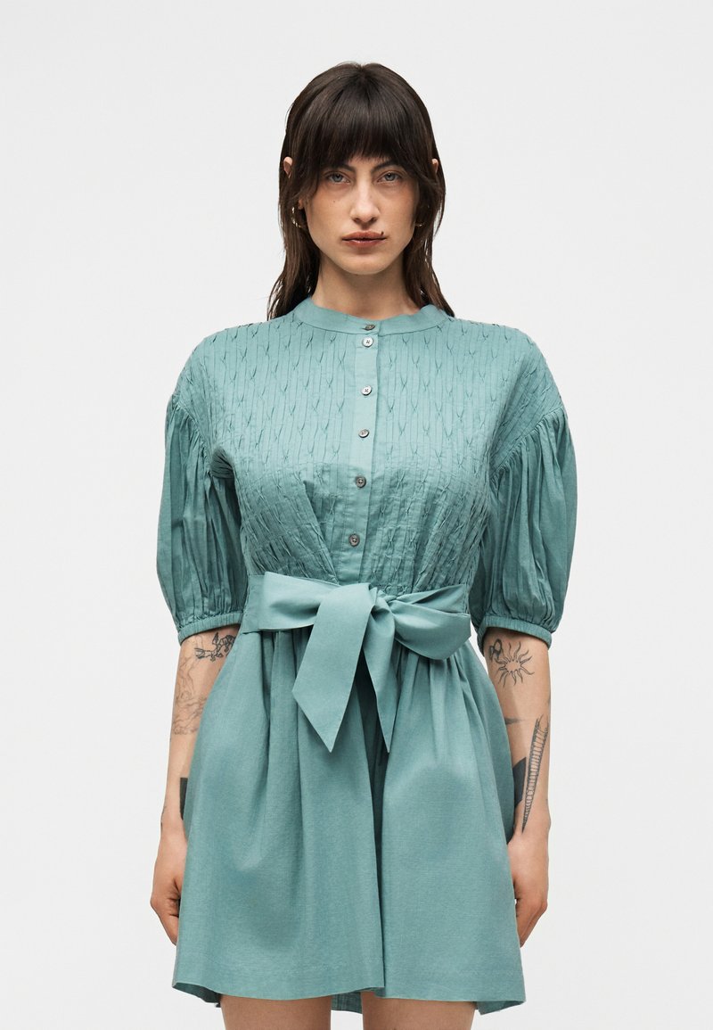 Γυναίκα που φοράει ένα μπλε-πράσινο φόρεμα με κουμπιά, φουσκωτά μανίκια και φαρδιά ζώνη στη μέση, που δείχνει τατουάζ σε και τα δύο πήχεις, στέκεται μπροστά σε απλό φόντο.