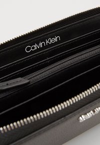 Juodas odinis piniginė su užtrauktuku, turintis kelis skyrius ir įspaustą "Calvin Klein" logotipą viduje. Sidabriniai aksesuarai.
