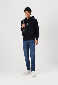 Hanorac bleu marin cu logo, blugi din denim și teneși albi. Persoana poartă o geantă de umăr neagră cu o curea branduită.