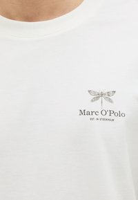 Λευκό μπλουζάκι με μικρό γκρι γραφικό λιβελούλης πάνω από μαύρο κείμενο "Marc O'Polo EST. IN STOCKHOLM" στο στήθος.