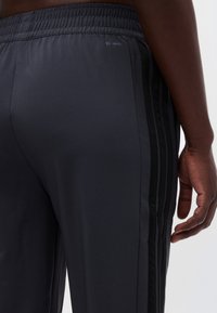 Κοντινό πλάνο ενός ατόμου που φοράει μαύρο αθλητικό παντελόνι με ελαστική μέση και διακριτικές λωρίδες στο πλάι, το χέρι του χαλαρό δίπλα στο σώμα.