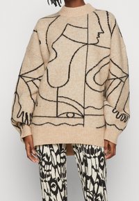 Beige oversized Pullover mit schwarzen abstrakten Linienmustern. Rippstrickkragen und -bündchen. Kombiniert mit gemusterten schwarz-weißen Hosen.