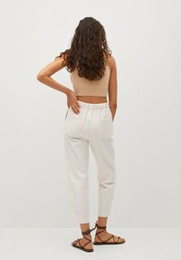 Beżowy krótki top, białe spodnie z elastycznym paskiem i kieszeniami oraz brązowe sandały na ramiączkach. Kręcone włosy swobodnie opadają na plecy.