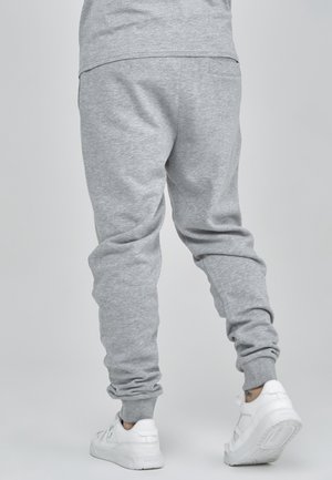 Person iført lysegrå joggingbukser og hvide sneakers, stående på en ensfarvet lysegrå baggrund, set bagfra.