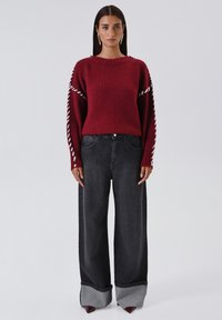 Donna con capelli scuri e lisci indossa un maglione a maglia bordeaux con cuciture bianche sulle maniche, un paio di jeans neri a gamba larga con risvolti e scarpe a punta.