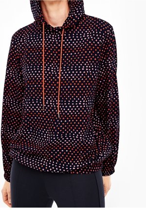 Kobieta ubrana w czarną bluzę z kapturem w czerwone i różowe rozsypane groszki, z pomarańczowymi sznureczkami i czarnymi dopasowanymi spodniami.