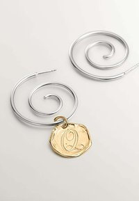 Pendientes en espiral de plata con un colgante de oro texturizado que presenta una 'Q' en relieve en el centro. Diseño de gancho simple, estética minimalista.