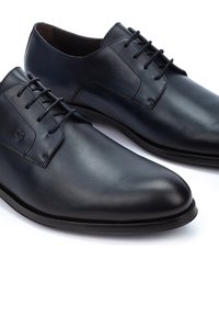 Scarpe da uomo in pelle nera con superficie liscia, punta rotonda e chiusura con lacci. Presentano un branding discreto e una suola nera robusta.