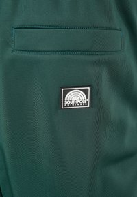 Zöld szövet zseb fekete címkével, amelyen egy fehér szivárvány logó és az alján a "SOUTHPACK ORIGINALS" felirat található. Simább textúra.