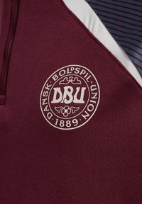 Bordeauxfarbener Sporttrikot mit strukturierter Oberfläche, das ein rundes weißes Emblem mit "DBU" und "DANSK BOLDSPIL UNION 1889" in fetter Schrift zeigt.