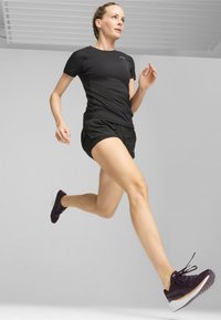 Top atlético negro de manga corta, pantalones cortos negros y zapatillas de correr moradas con suelas blancas; mujer en medio de una zancada contra un fondo gris.