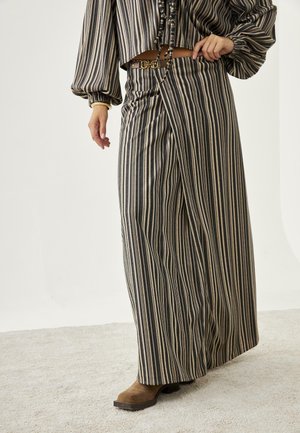 Mujer con pantalones de pierna ancha de rayas verticales y cintura alta, con un cinturón marrón y un top a juego de manga abullonada, de pie sobre una alfombra.