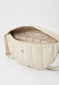 Beige quiltad axelväska med en slät yta, en logopatch och ett zippered öppning som avslöjar ett mjukt, fodrat innandöme.