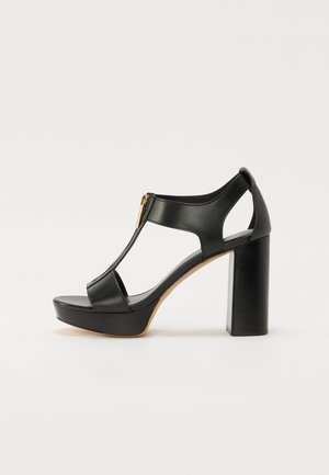 Sandale en cuir noir à talon haut, bout ouvert, design à bride en T, détail de fermeture éclair dorée et talon bloc épais sur fond blanc.
