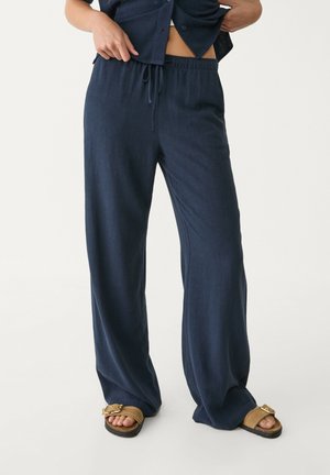Personne portant un pantalon ample bleu marine avec taille élastique et cordon de serrage, associé à des sandales à enfiler couleur sable sur un fond blanc.