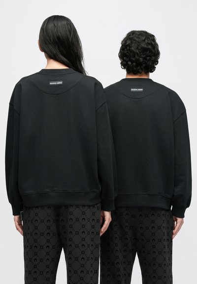 Melni džemperi ar apaļu kaklu, platu piedurknēm un ribotām aprocēm, kakla izgriezumā ar etiķeti; kombinēti ar melnām biksēm ar rakstu.