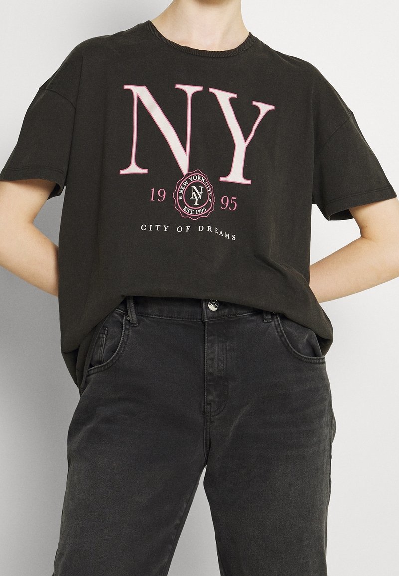 Personne portant un t-shirt noir court avec l'inscription "NY 1995 City of Dreams" en lettres roses et blanches, associé à un jean noir taille haute.
