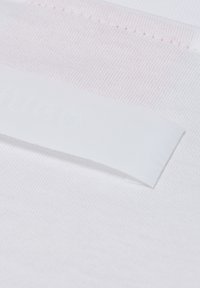 Wit katoenen stof met een gestructureerd oppervlak; bevat een lichtroze streep en een vlak wit label met reliëf tekst.