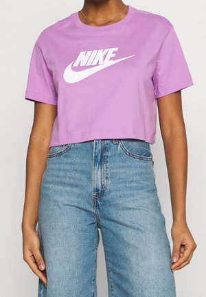 Persoon in een paars, kort Nike T-shirt en high-waisted blauwe jeans, met ontspannen handen langs de zijden tegen een effen achtergrond.