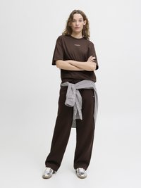 Bruine T-shirt met korte mouwen en een wit logo, gecombineerd met losse bruine broek en een geblokt overhemd om de taille geknoopt. Zilveren sneakers.