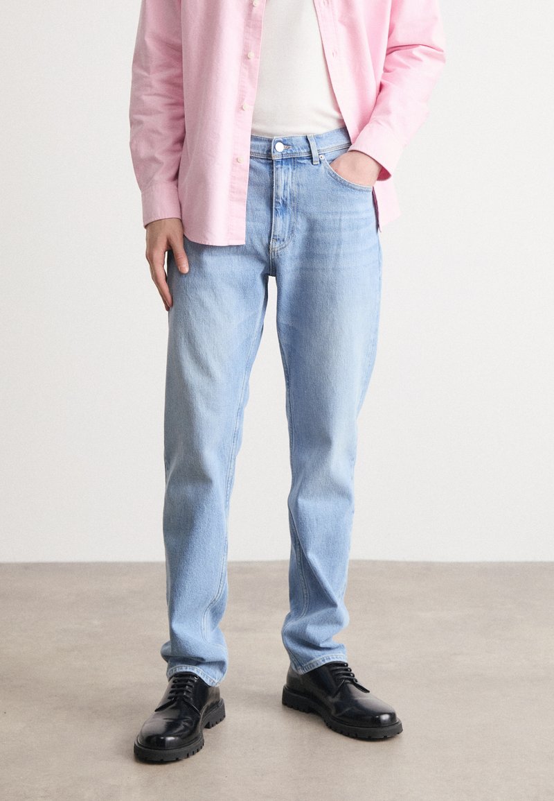 Lys blå jeans med rett passform, denimmateriale, kontrasterende sømmer og frontlommer. Kombinert med en rosa skjorte med knapper og svarte sko.