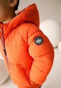 Doudoune orange vif avec fermeture éclair à l'avant, capuche attachée et un patch rond avec un logo représentant des montagnes sur l'épaule gauche.