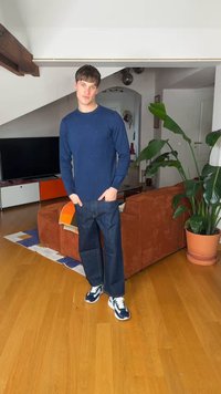 Mladý muž v modrém svetru a džínách stojí v obývacím pokoji s oranžovou pohovkou, velkou květinou v květináči a dřevěnou podlahou.