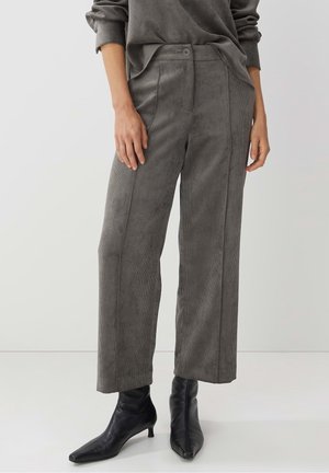 Nainen, joka seisoo vaaleaa yksiväristä taustaa vasten, pukeutuneena harmaisiin, leveälahkeisiin, corduroy-housuihin ja mustiin, kärjestä teräviin nilkkureihin.