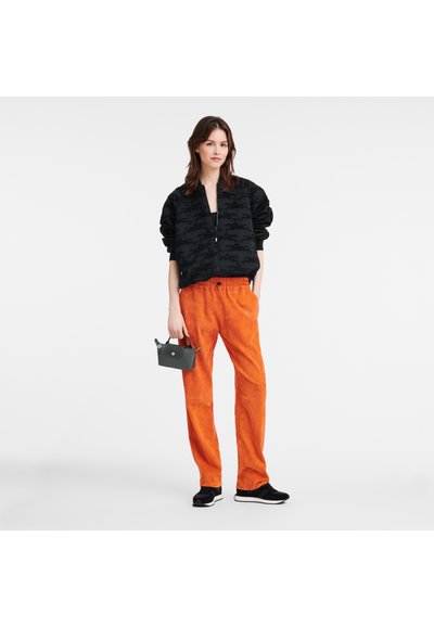 Black bomber jacket with textured pattern, bright orange suede pants, gray handbag, and black sneakers. Casual outfit with bold color contrast.